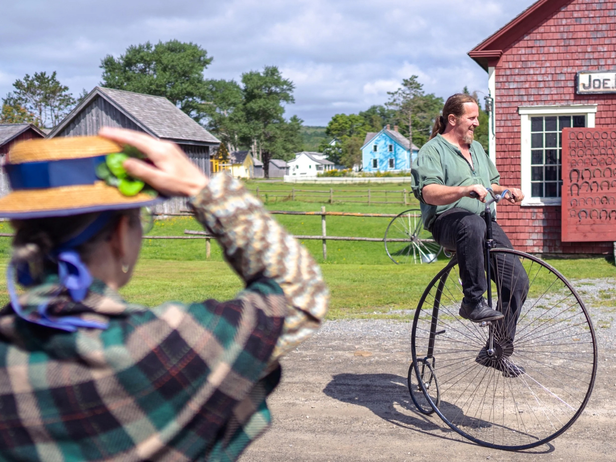 Costumed interpreter, a man, riding a penny-farthing past the blacksmith shop.