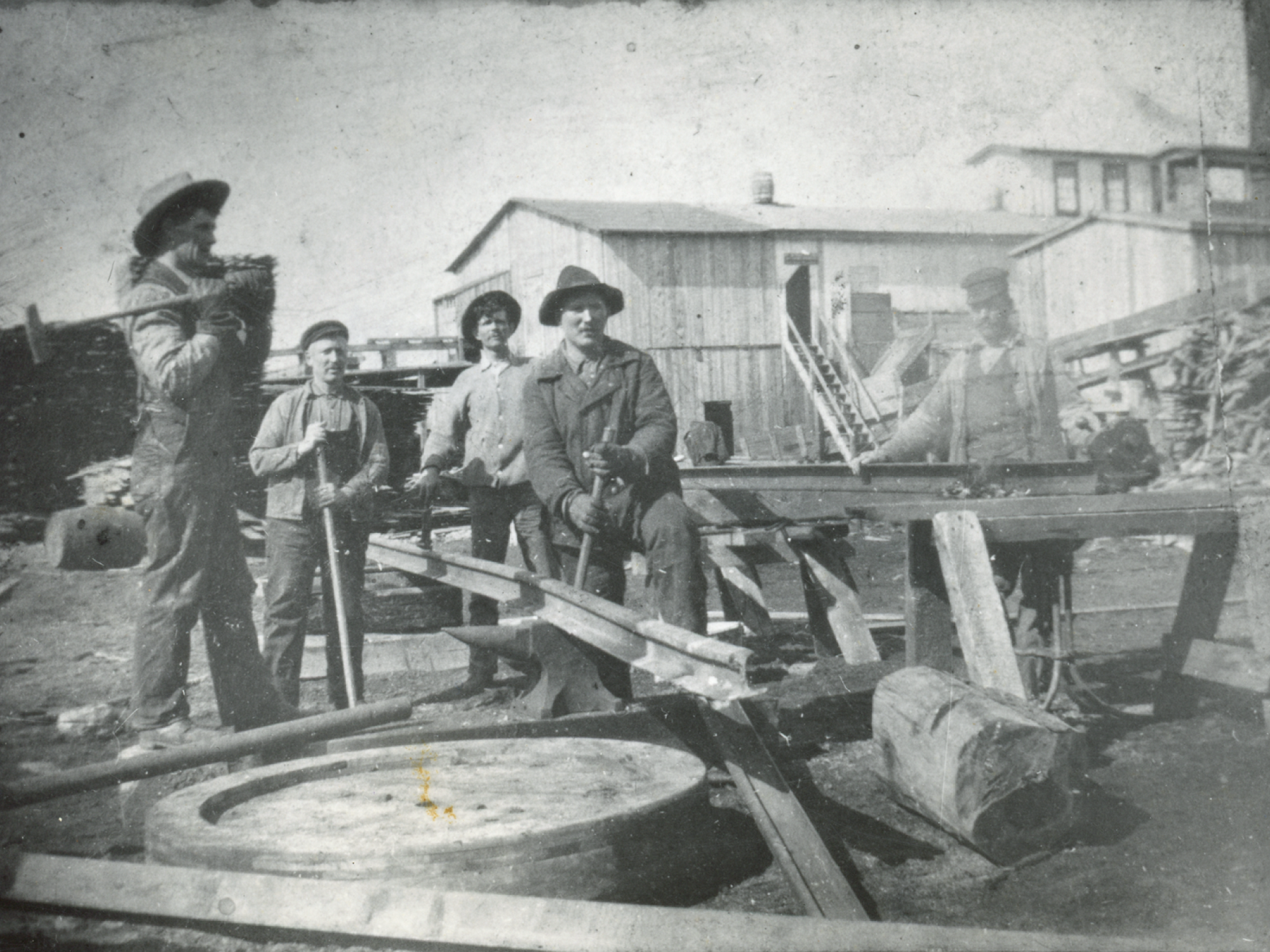Gemini said A vintage black and white photograph showing five men posed at an industrial outdoor work site, likely a mill or railway yard.  The men are dressed in early 20th-century work clothing, including wide-brimmed hats, flat caps, and overalls. They are positioned around several long metal rails, a large anvil, and wooden sawhorses. 