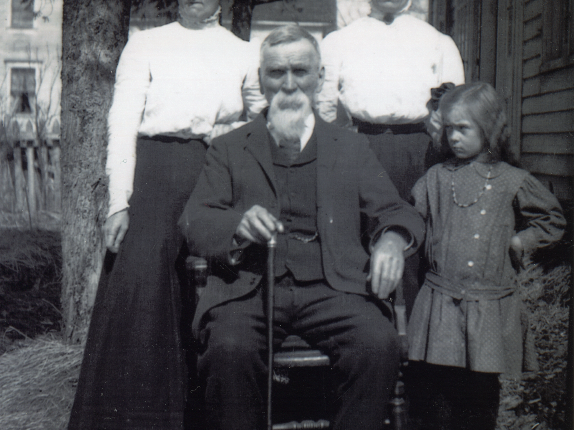 A vintage black and white photograph from the early 20th century featuring a family portrait outdoors.  In the center, an elderly man with a long white beard and mustache sits in a chair, holding a walking cane between his knees. Behind him stand two women with their hair styled in voluminous updos, wearing high-necked white blouses and long dark skirts. To the right, a young girl in a dark, patterned dropped-waist dress and a necklace stands with a somber expression. The group is posed on a grassy area.