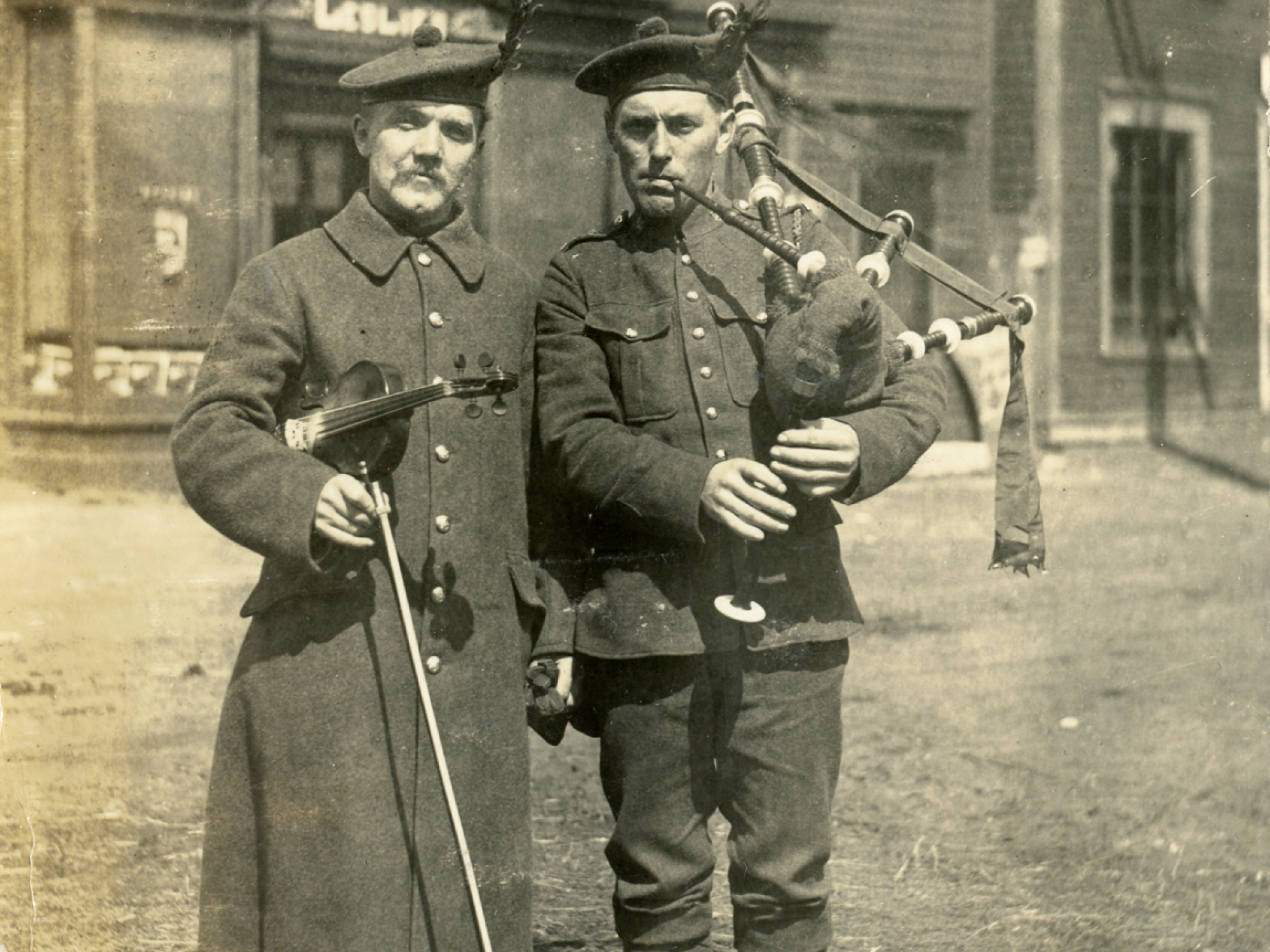 A sepia-toned vintage photograph from April 1916 featuring two men in Highland-style military uniforms posing with musical instruments.  The man on the left wears a long, heavy greatcoat and a Glengarry bonnet with a feather, holding a fiddle and bow. The man on the right wears a military tunic, puttees (leg wraps), and a similar feathered bonnet while playing the bagpipes.