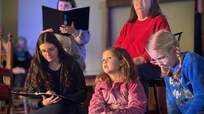 An older woman in a vibrant red sweater sits on a chair, looking upward with a focused expression. In front of her, three young people sit on the floor: a teenage girl on the left looking down at an open binder in her lap, a young girl in the center wearing a pink jacket looking upward, and another young girl on the right in a blue sweatshirt looking downward. In the background, a person holds a large black binder open, and another blurred figure sits further back on the left.