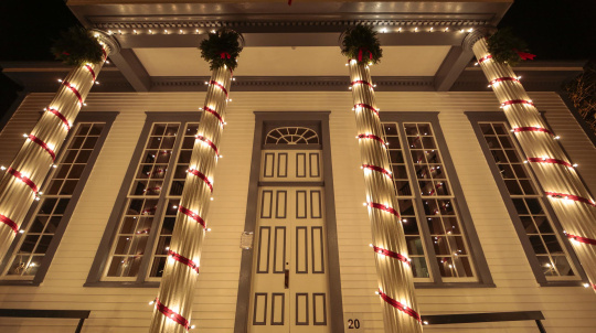 A white, two-story building with tall columns decorated for Christmas at night. The four front columns are wrapped in bright white string lights and a twisting red ribbon. Each column is topped with a green wreath. The building's front door is centered and flanked by two large, multi-paned windows on each side.